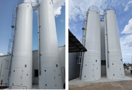 Instalação de dois silos industriais rígidos brancos lado a lado, equipados com escadas, passarelas e tubulação, em frente a um edifício sob um céu azul.