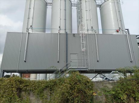 Quatro silos industriais rígidos brancos alinhados em uma plataforma metálica com escada de segurança e tubulação, em frente a uma fachada com painéis cinza.