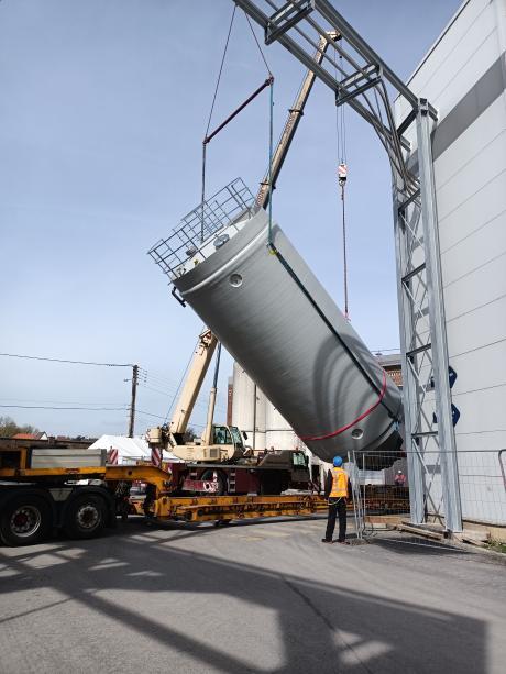 Operário com colete de alta visibilidade orientando a instalação de um silo industrial rígido inclinado por um guindaste móvel montado em um caminhão plataforma no local de produção.