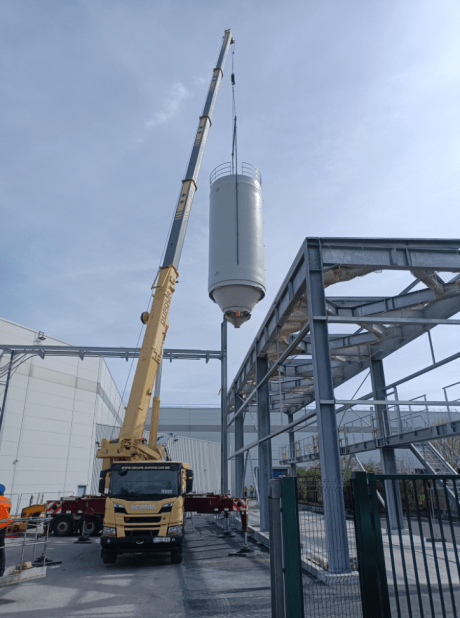 Guindaste móvel posicionando um silo industrial rígido branco sobre uma estrutura metálica externa em uma unidade de produção.