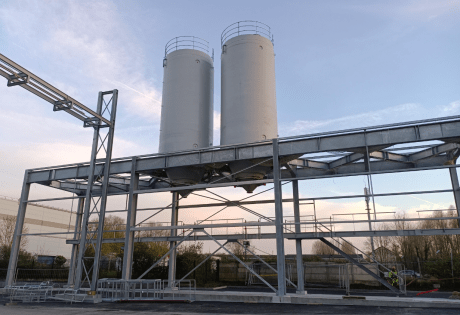 Dois silos industriais rígidos brancos instalados sobre uma estrutura metálica externa com passarela e estrutura de acesso, fotografados ao entardecer em um local de produção.