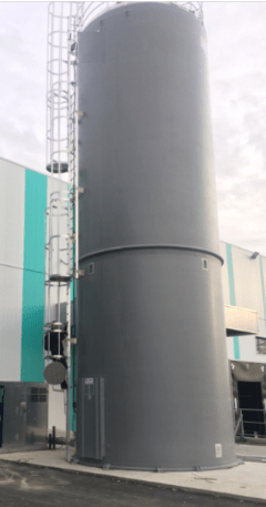 Silo rígido industrial cinza antracite equipado com escada e guarda-corpo em aço, instalado em frente a uma fachada com painéis brancos e turquesa.