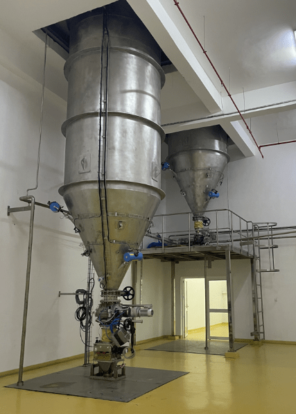 Dois silos rígidos em aço inoxidável instalados no interior sobre uma plataforma metálica com válvulas e tubagem para alimentação e regulação de um processo industrial.