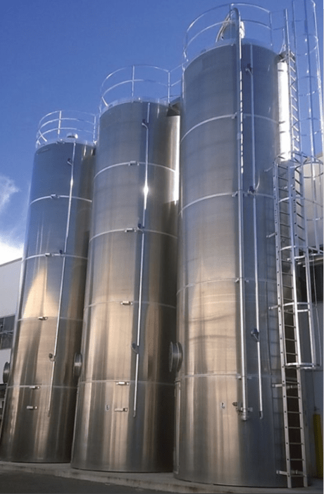 Três silos industriais rígidos em aço inoxidável brilhante, equipados com escadas e corrimãos, instalados no exterior sob um céu azul.